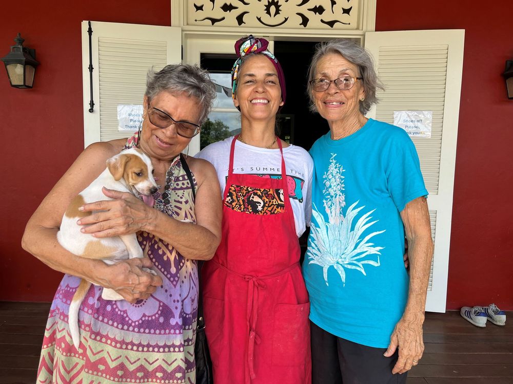 Three women smiling outdoors, one holding a small dog, another wearing a red apron, in front of a red building.