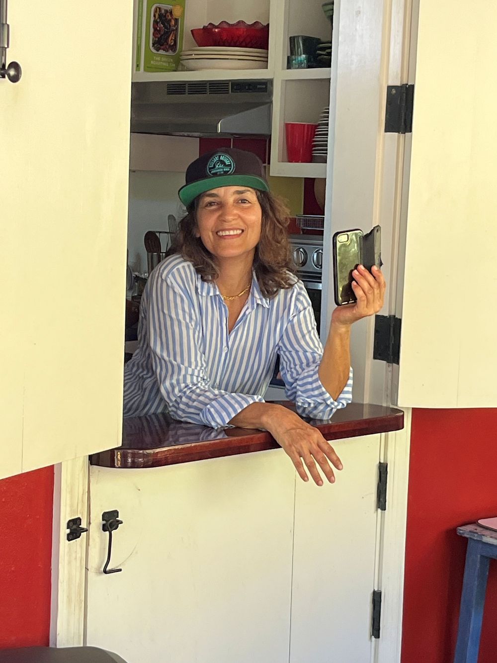 Smiling woman in green cap and striped shirt leaning out of a Dutch door, holding a phone.