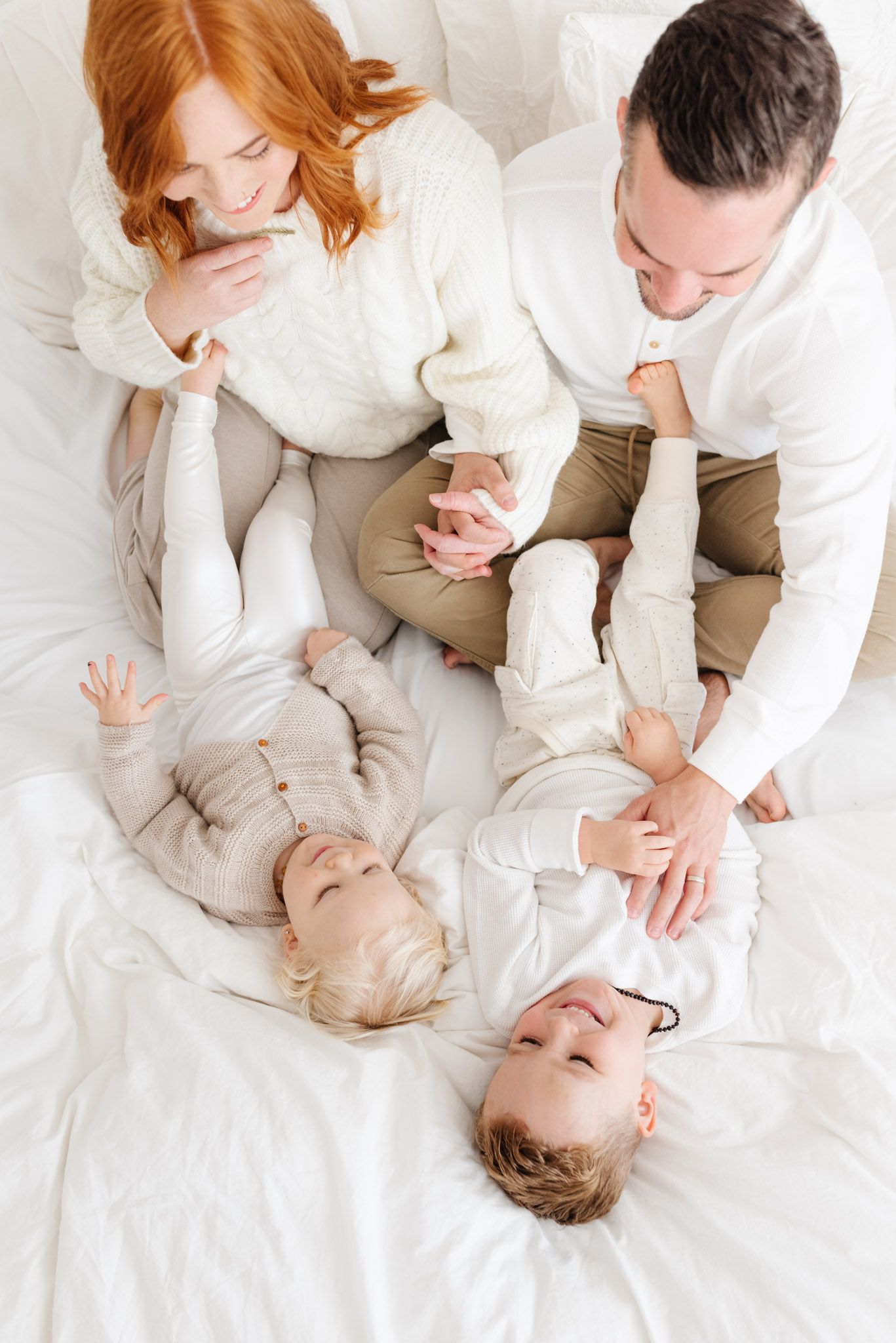 A happy family with two toddlers playing on a white bed, parents in cream sweaters enjoying a joyful moment.