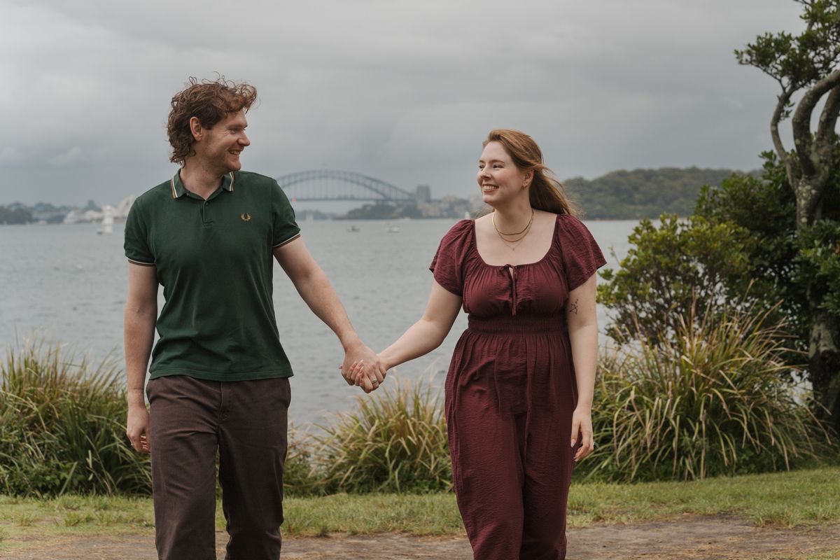 Couple holding hands and smiling at each other near Sydney Harbour Bridge on a cloudy day during engagement photoshoot in sydney Milk Beach