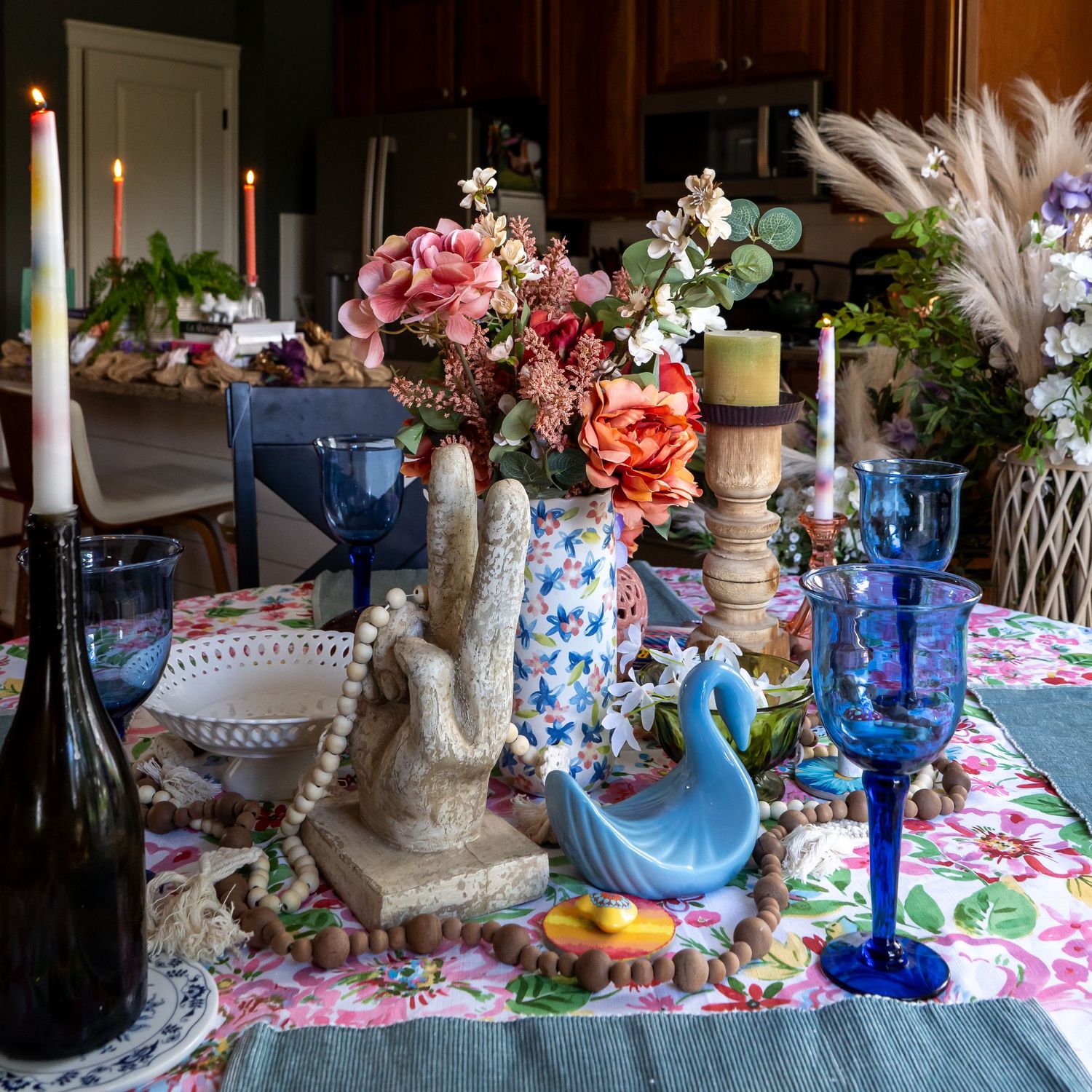 Colorful dining table with floral centerpiece, blue glassware, candles, and decorative swan figurine on a floral tablecloth.