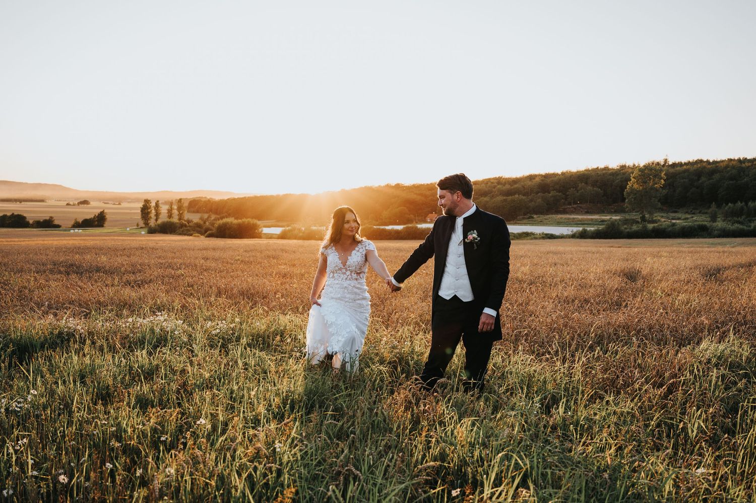 Bride and groom holding hands walking through golden wheat field at sunset on their wedding day.