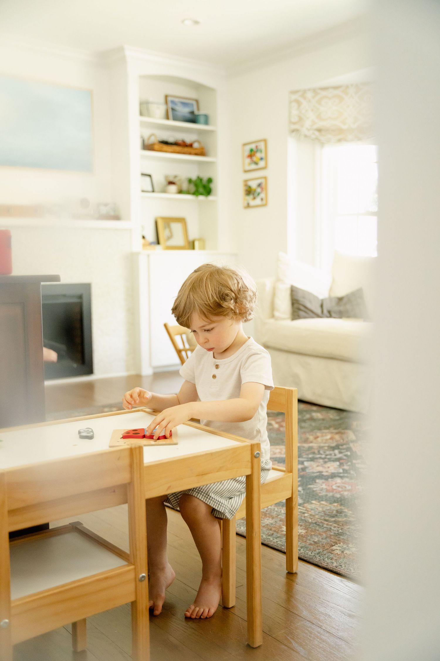 Young toddler with curly hair sits at a small wooden table playing with red toy in a bright, cozy living room.