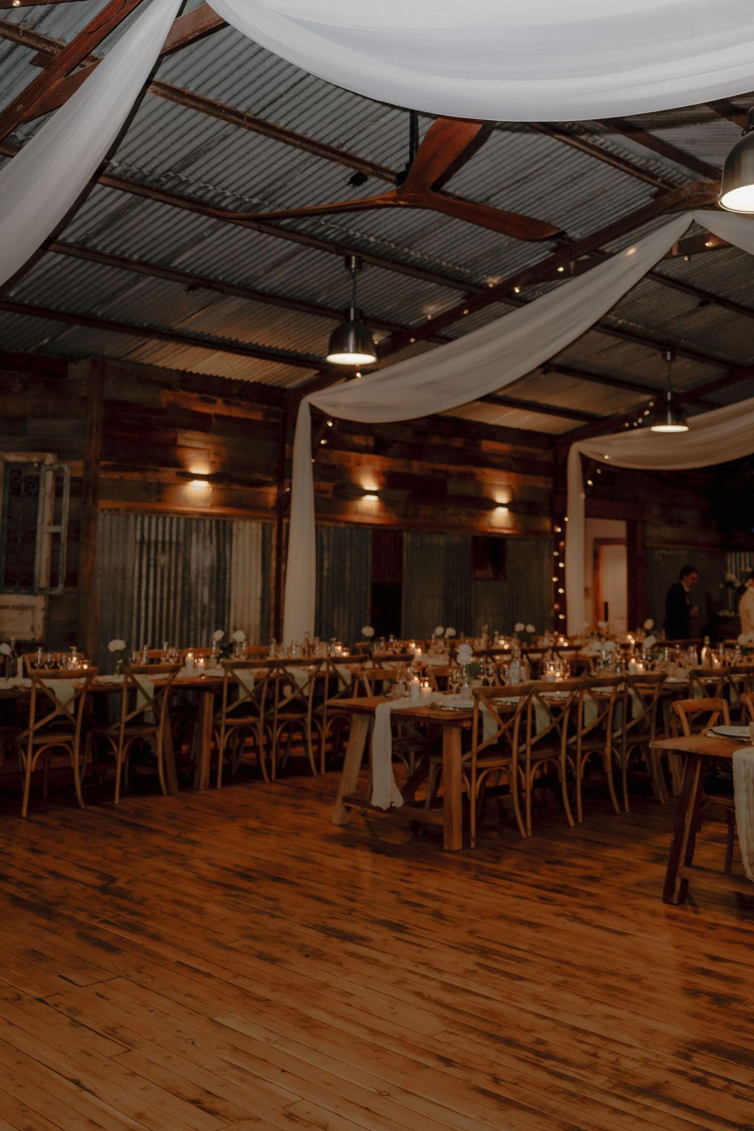 Rustic barn wedding reception with long tables, candles, white draping, string lights, and warm wood floors.