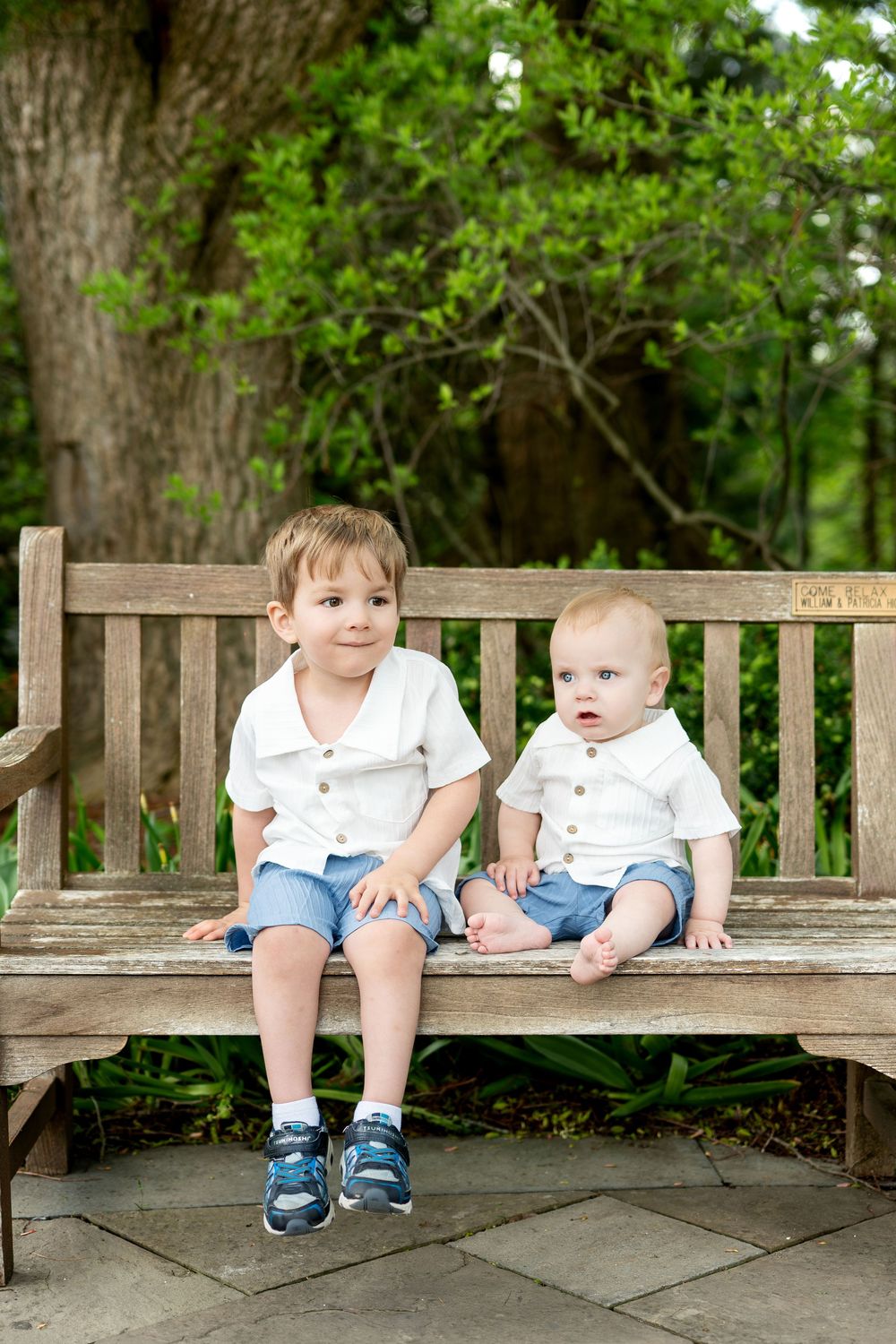 Two young brothers in matching white shirts and blue shorts sitting together on a wooden park bench.