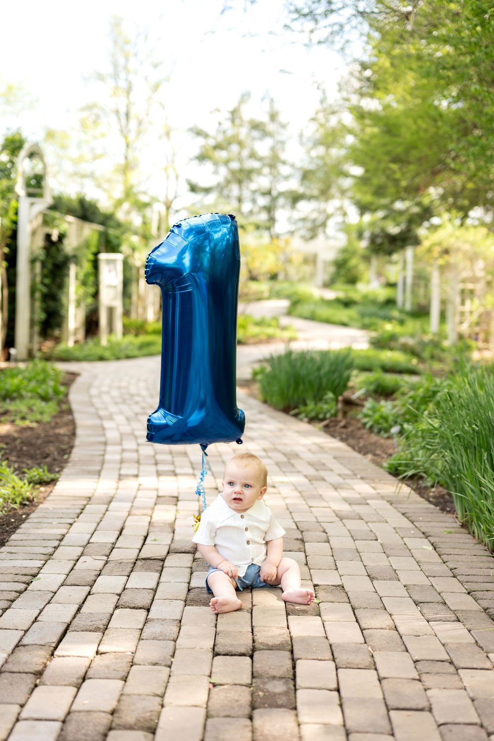 Baby sits on brick path holding blue number 1 birthday balloon.