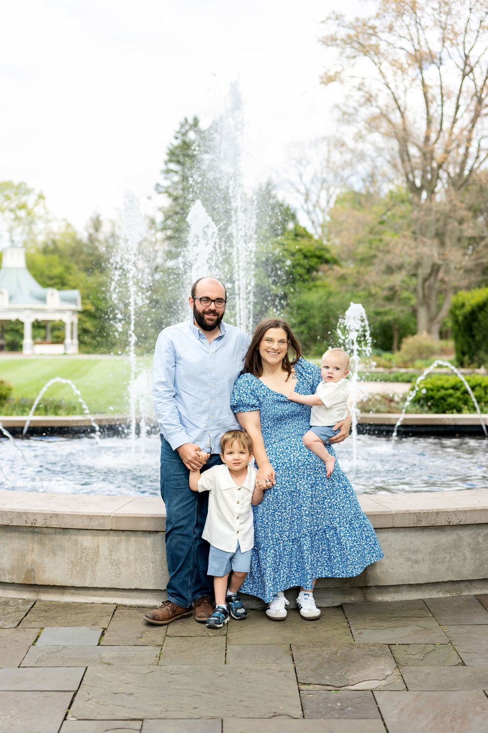 Smiling family of four with two young children posing before a beautiful outdoor fountain.