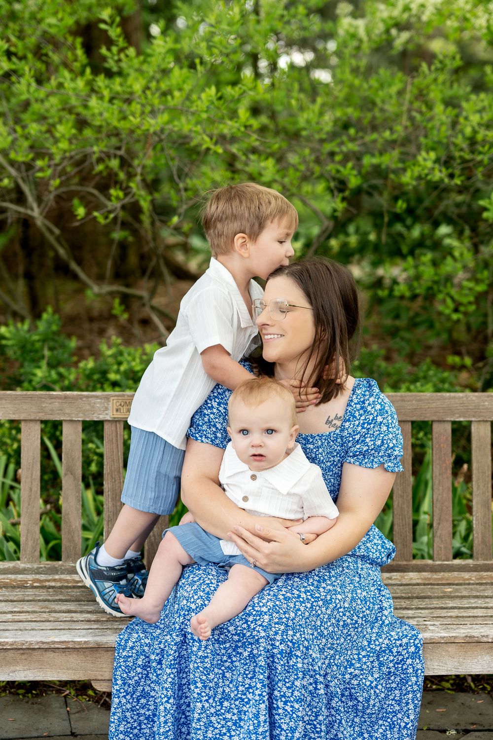 Mother in blue floral dress on garden bench laughs as toddler son whispers in her ear while she holds her baby.