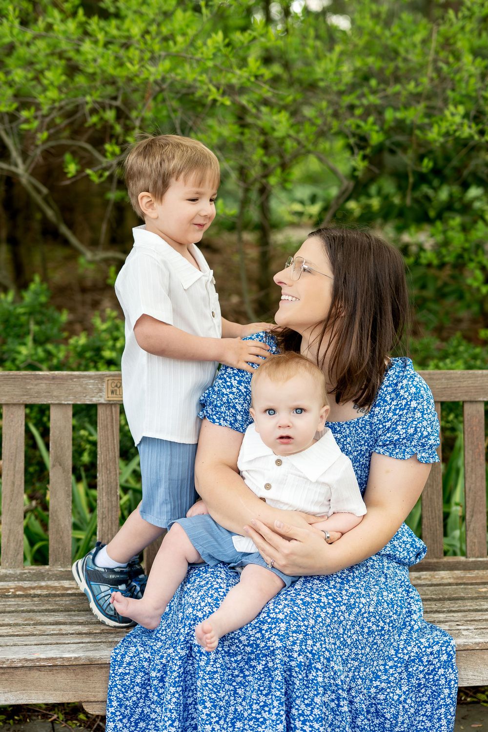 Smiling mother in blue floral dress sits on wooden bench with toddler son standing beside her and baby on her lap.