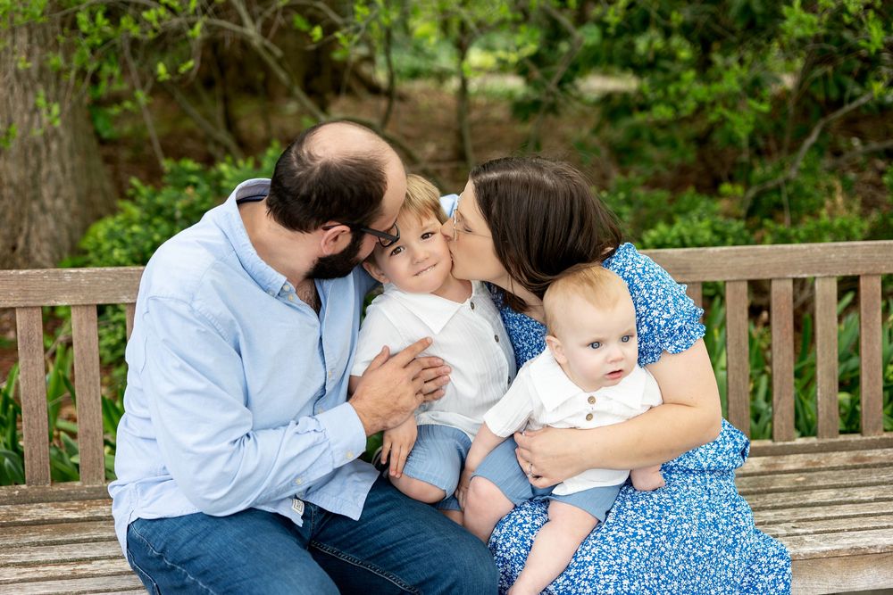Parents lovingly kiss their toddler son on the cheeks while mom holds baby on a wooden bench in a lush garden.