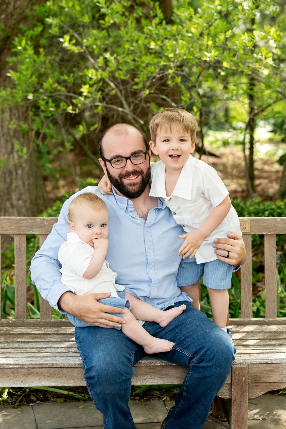 Smiling bearded father in glasses sits on wooden bench holding baby while excited toddler son stands beside him outdoors.