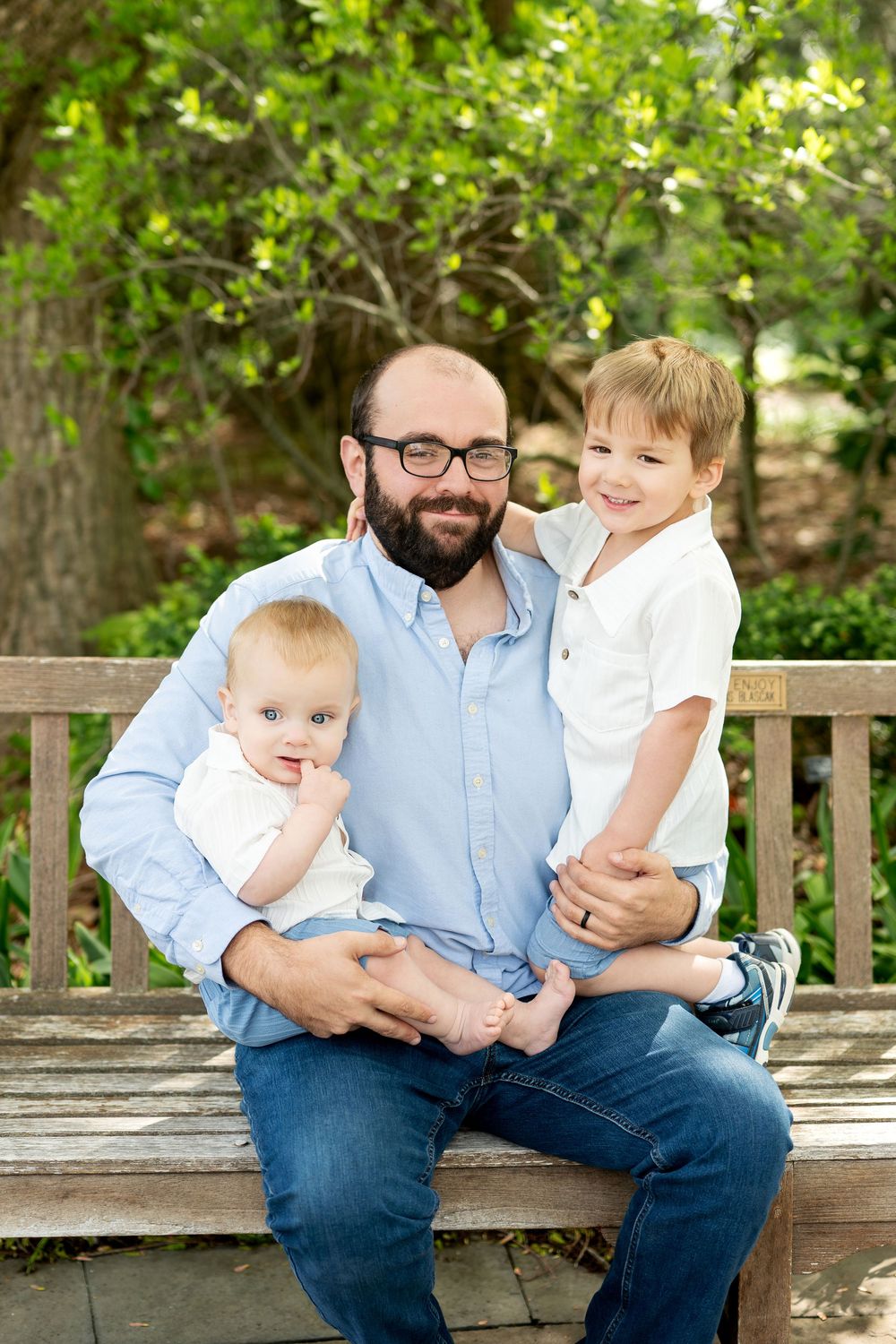 Bearded dad wearing glasses poses on a wooden garden bench with a baby and a smiling toddler son in white shirt.
