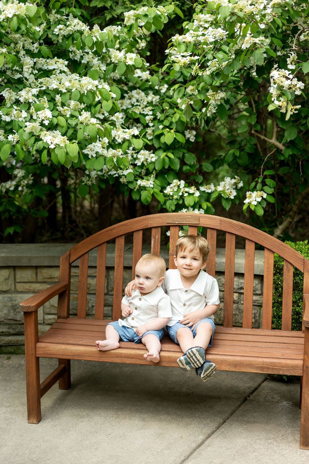 Two young brothers in white shirts sitting together on a wooden bench with flowering trees.