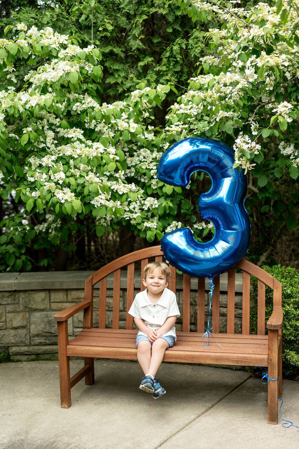 Smiling child sitting on wooden bench with large blue number 3 balloon among blooming trees.