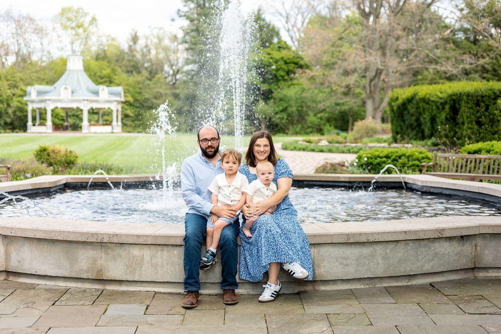 Family of four posing by a garden fountain, parents sitting with two young children in coordinating blue outfits.