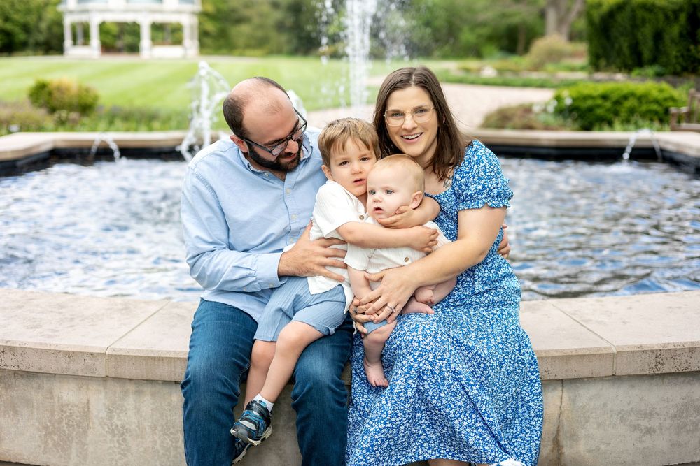 Smiling family of four sitting close together at a garden fountain, parents holding their two young sons.