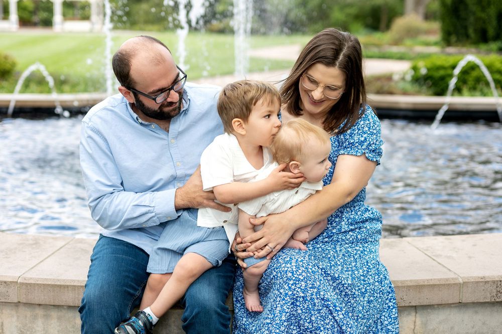 Tender family moment as toddler kisses baby sibling while parents watch lovingly by a park fountain.