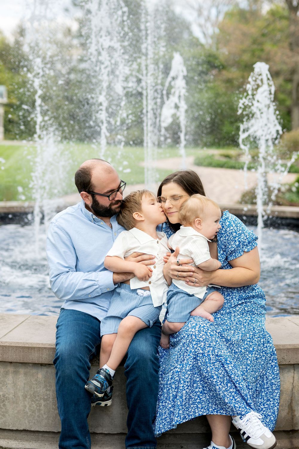 Loving family of four sharing kisses and cuddles in front of a cascading park fountain outdoors.