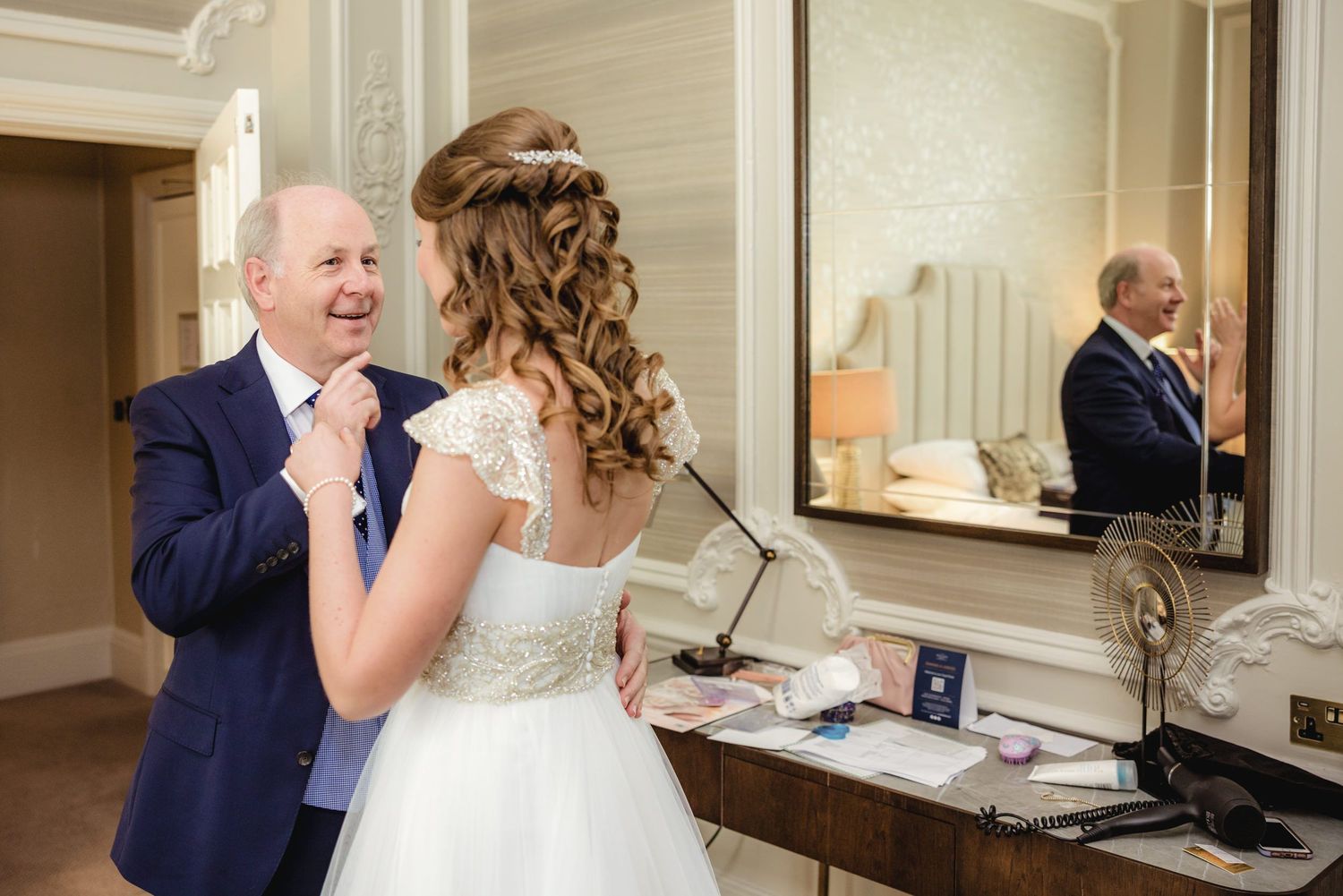 Bride in white wedding dress adjusting groom's navy tie, smiling together near mirror in elegant venue.