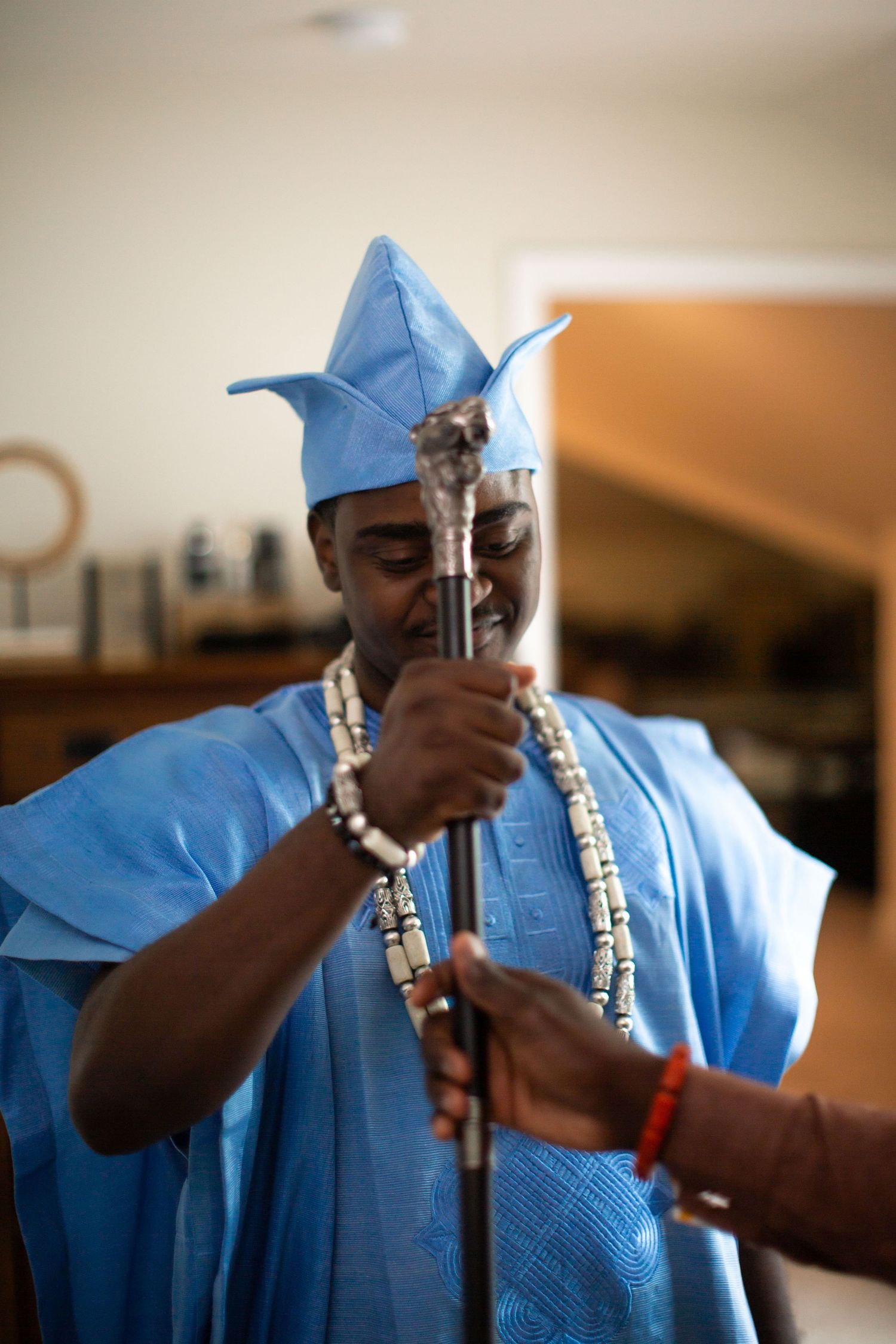 Man in blue Nigerian agbada attire and fila cap holding a ceremonial staff with beaded necklaces.