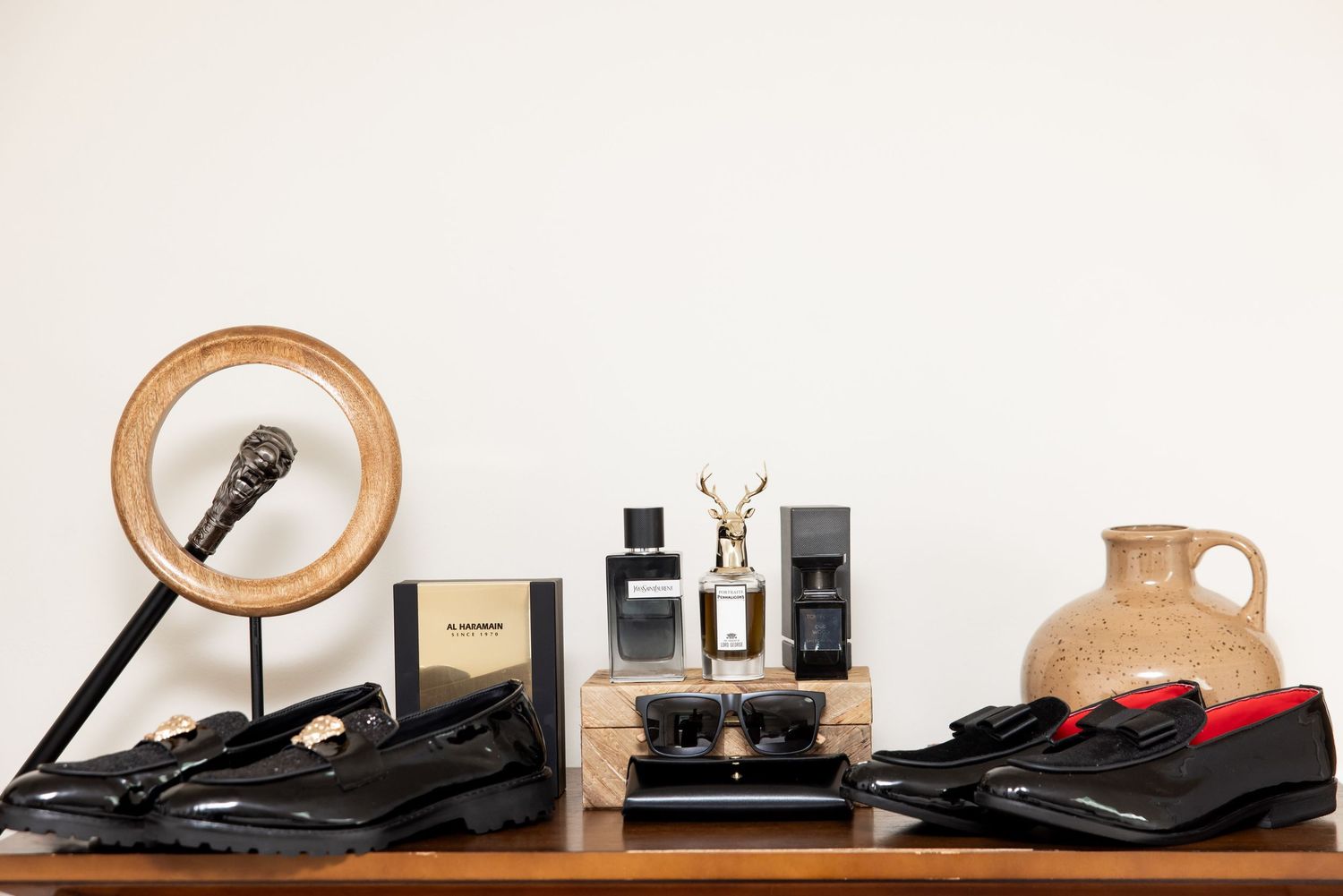 Black loafers and dress shoes displayed on a wooden shelf with cologne bottles, sunglasses, and decorative objects.