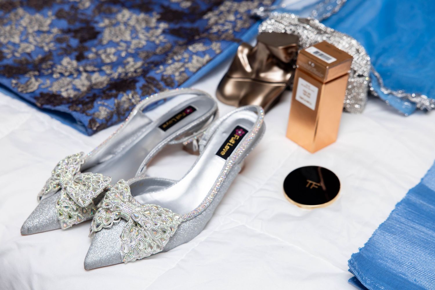 Silver embellished bow heels with perfume bottle, compact mirror, and blue fabric on white background.