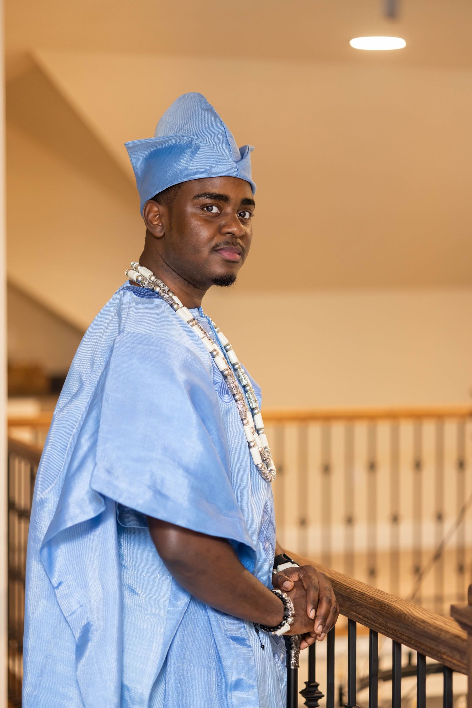 Young man in traditional blue African attire and matching cap, wearing beaded necklaces, standing by wooden staircase railing.