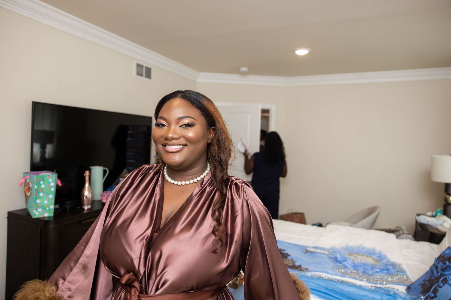 A smiling woman in a satin brown robe wearing pearl necklace stands in a bedroom during wedding preparations.