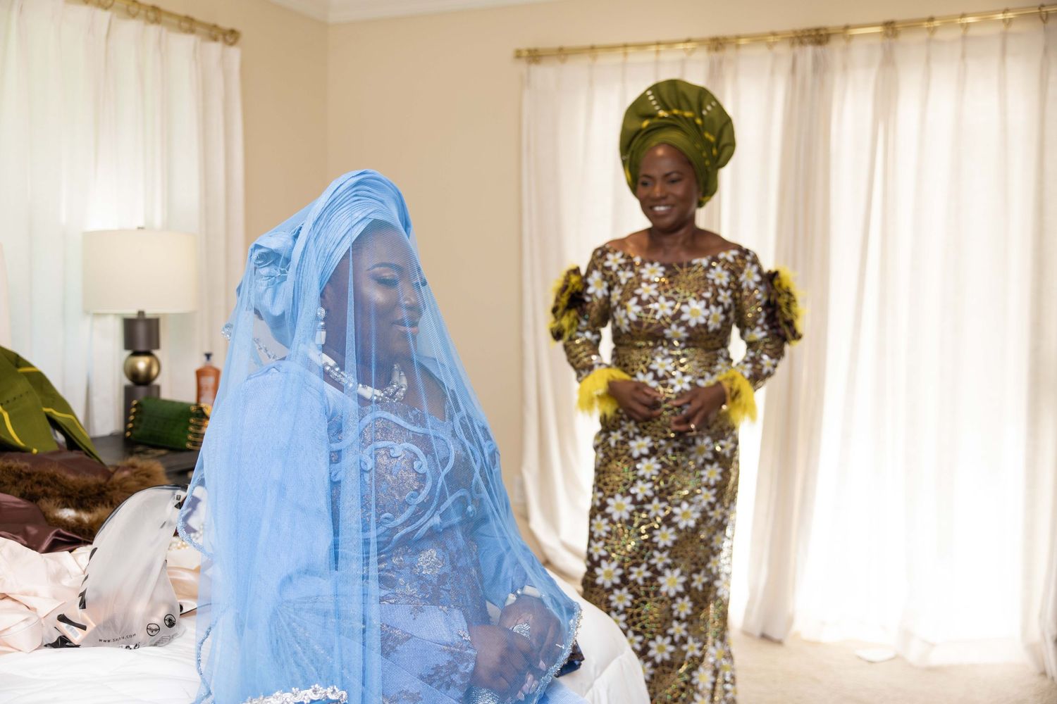 Two women in traditional African attire, one wearing a blue veil and the other a floral dress with green headwrap.