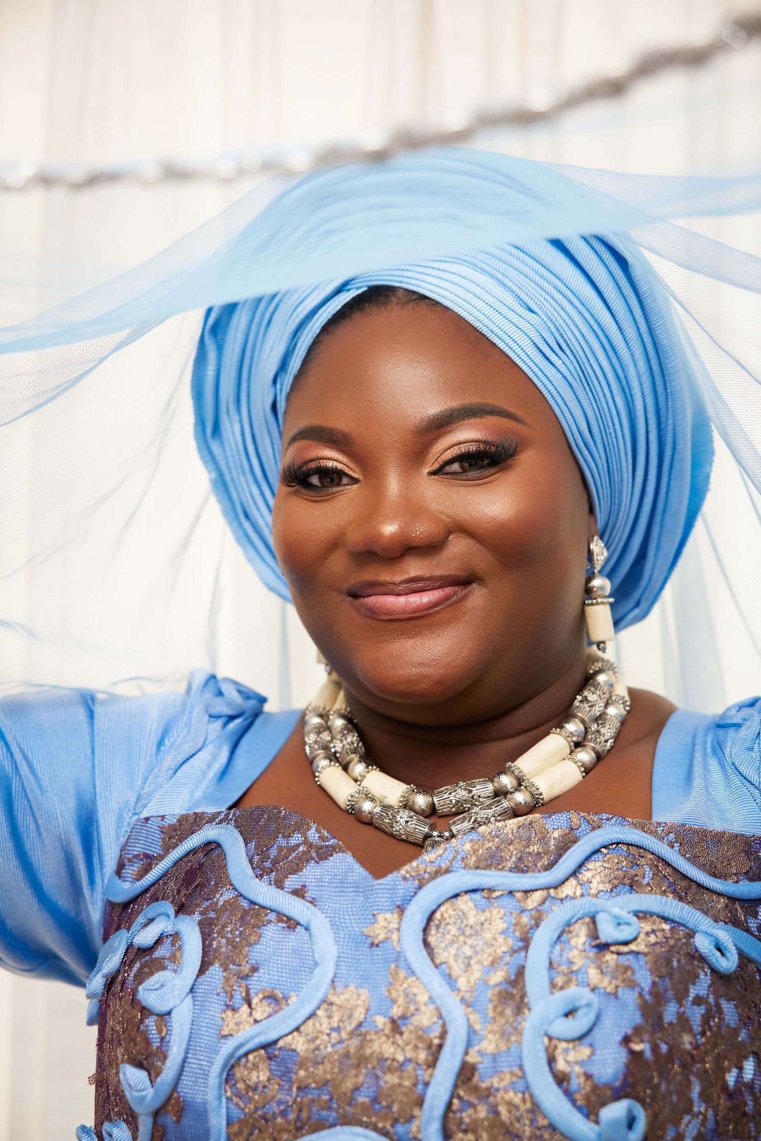 A smiling woman wearing a blue African gele headwrap, beaded necklace, and embroidered blue dress in a portrait photo.
