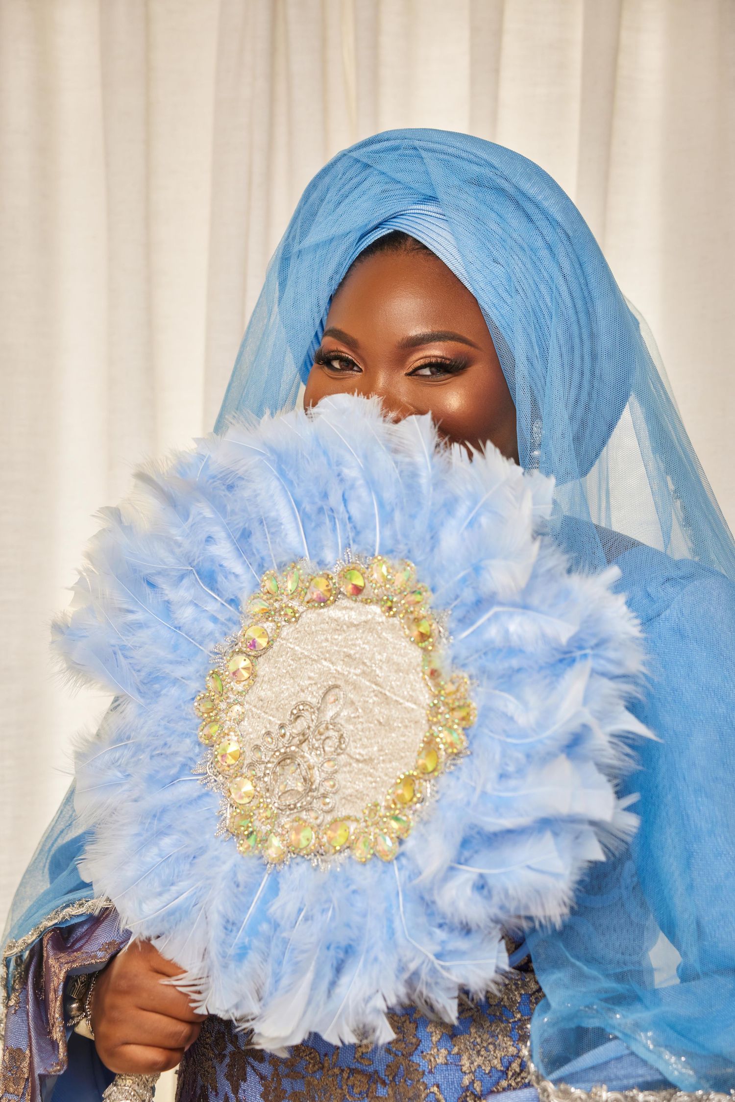 Woman in blue hijab and traditional attire holds decorative feather fan with jeweled embroidery at cultural celebration.