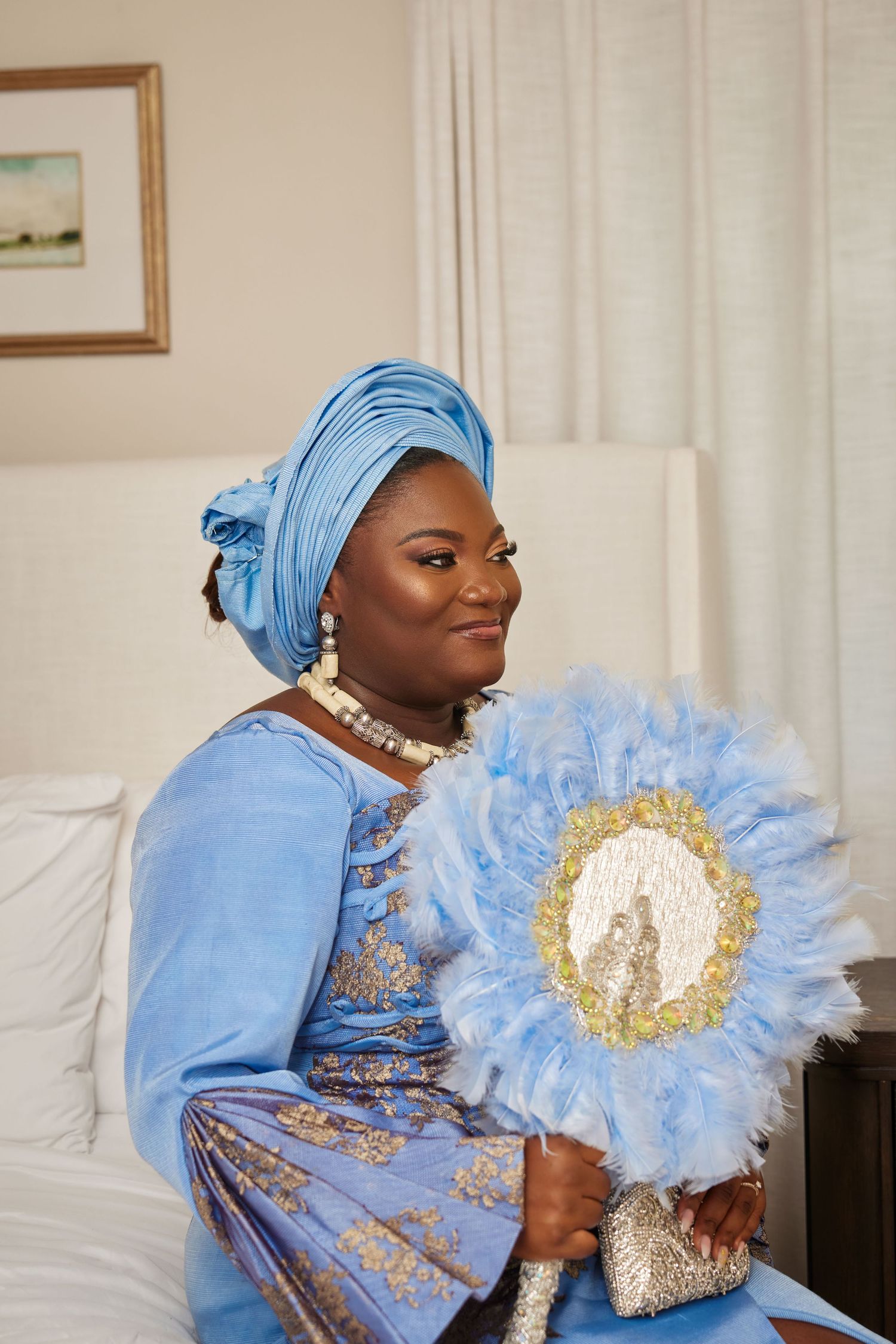 Woman in blue African traditional attire with gele headwrap, pearl necklace, and decorative feather fan.