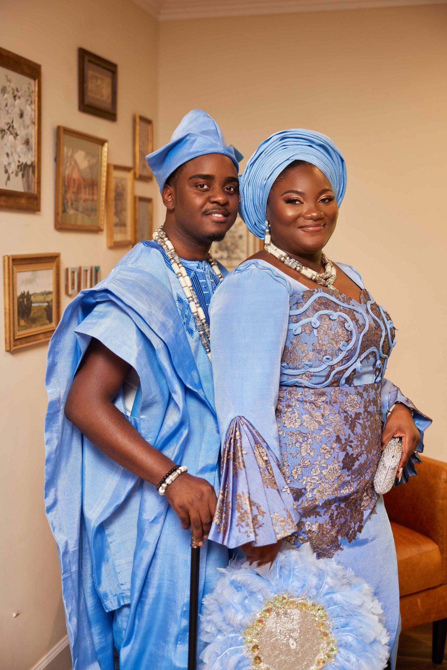 Couple in matching blue traditional Nigerian attire, gele headwrap, agbada, and lace dress, posing indoors.