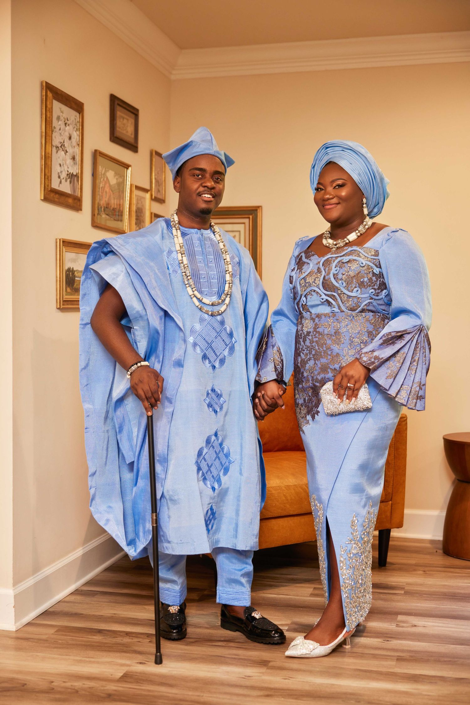 Couple in matching blue traditional African attire posing indoors, woman in gele headwrap, man holding a cane.