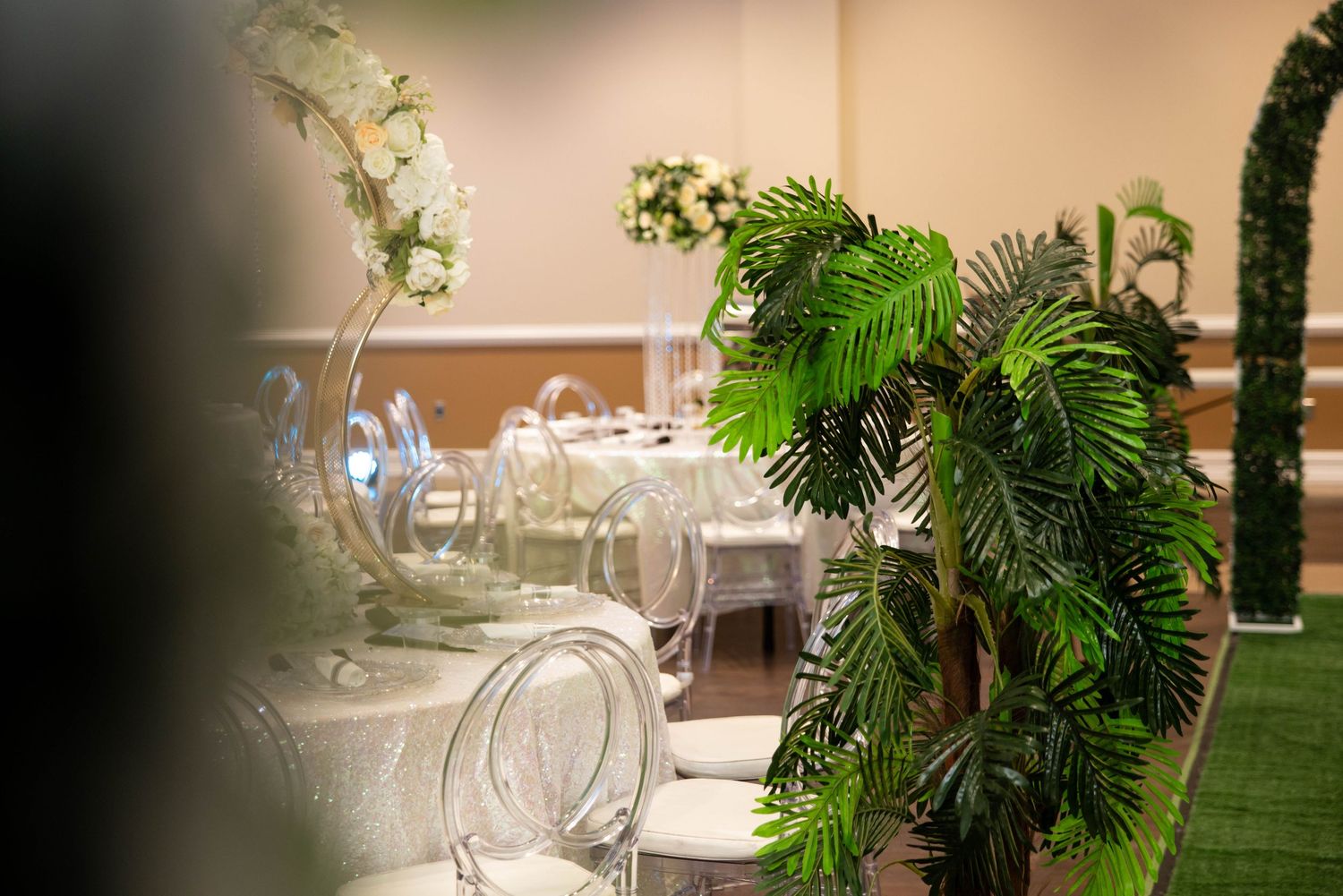 Elegant wedding reception venue with ghost chairs, white floral centerpieces, and lush tropical palm leaf decorations.