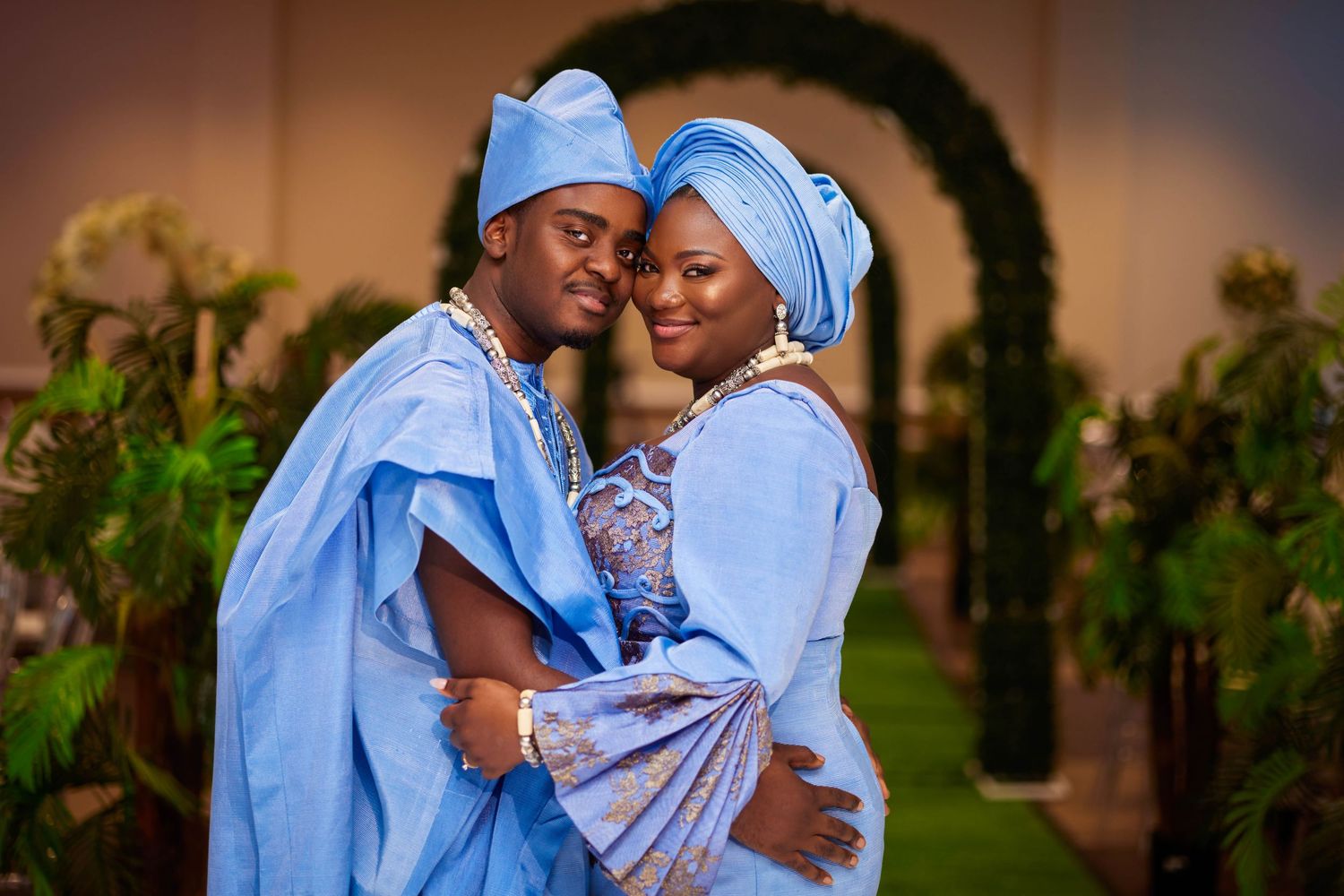 Couple in matching blue traditional Nigerian Yoruba attire smiling at a wedding ceremony with green floral arch backdrop.