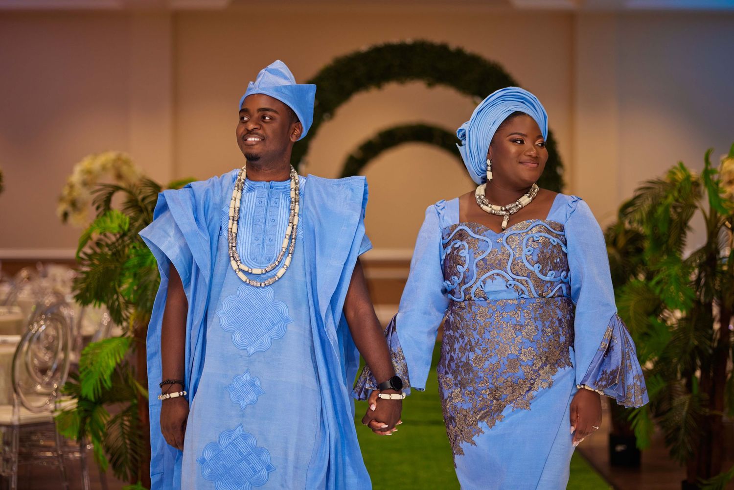 Couple in matching blue Nigerian traditional attire holding hands at an elegant wedding reception venue.