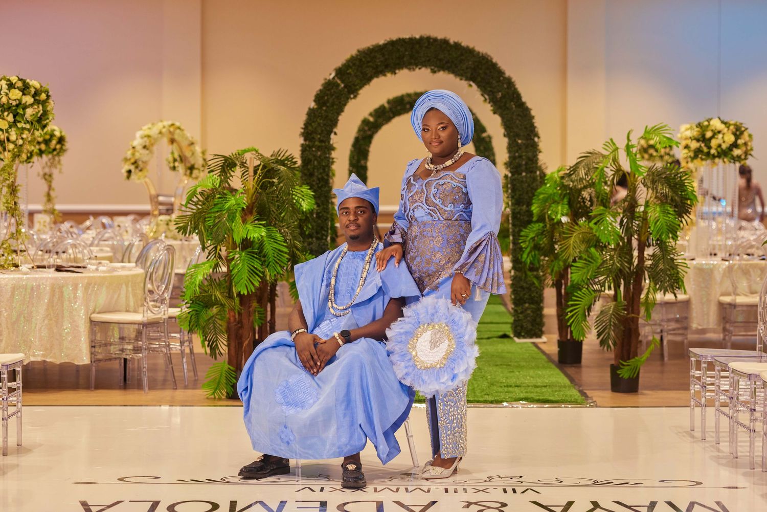 Couple in blue traditional Nigerian attire posing at an elegant wedding reception with floral arch backdrop.