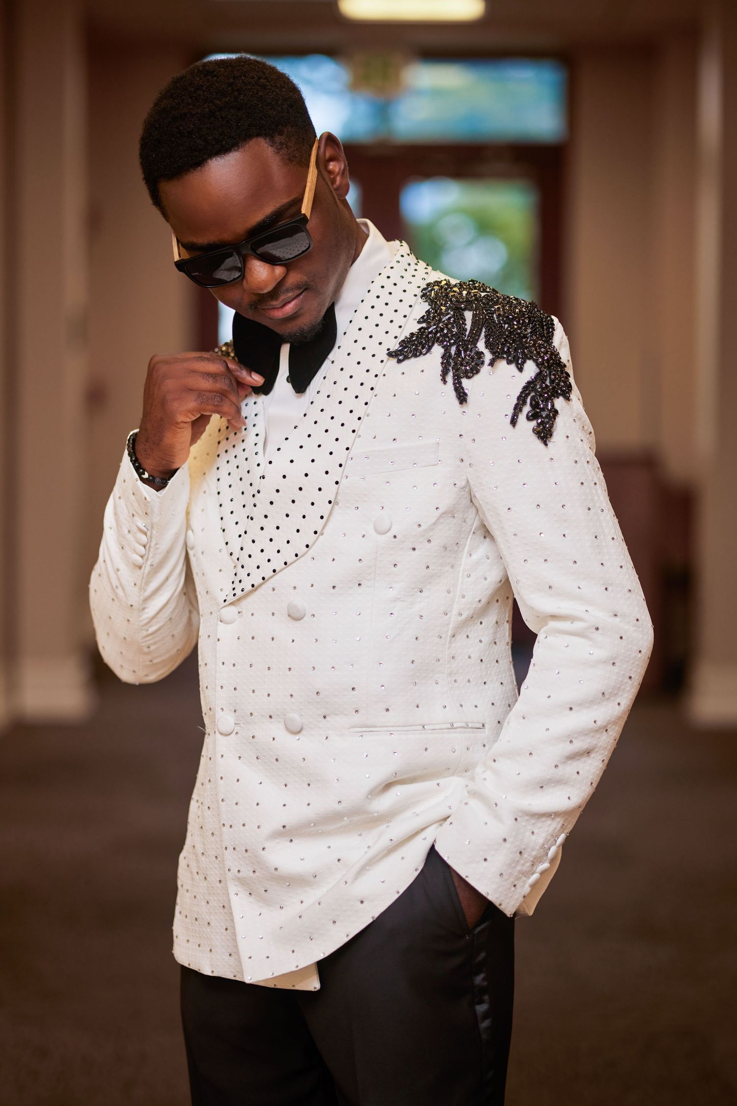 Stylish man in embellished white tuxedo jacket with black beading, sunglasses, and black pants posing indoors.