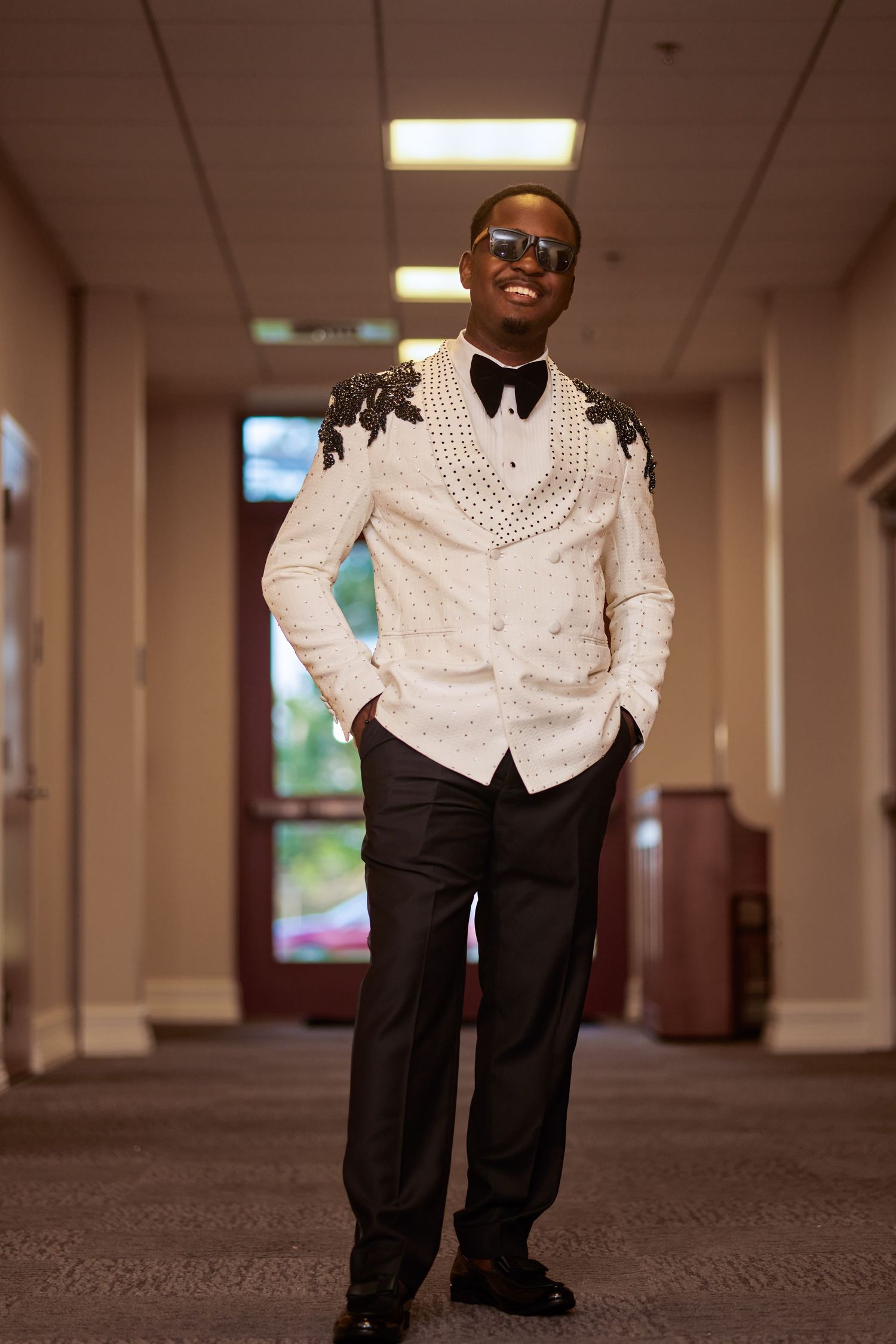 Stylish man in ornate white blazer with black floral embroidery, bow tie, sunglasses, and dark trousers posing in hallway.
