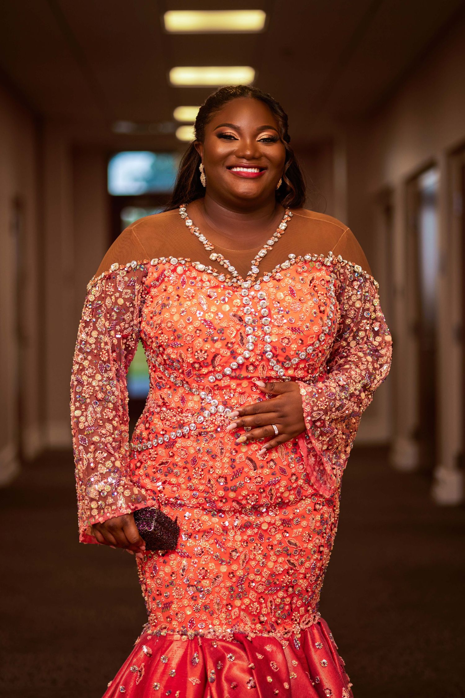 Woman in elegant beaded coral mermaid gown with sheer sleeves smiles in hallway at formal event.