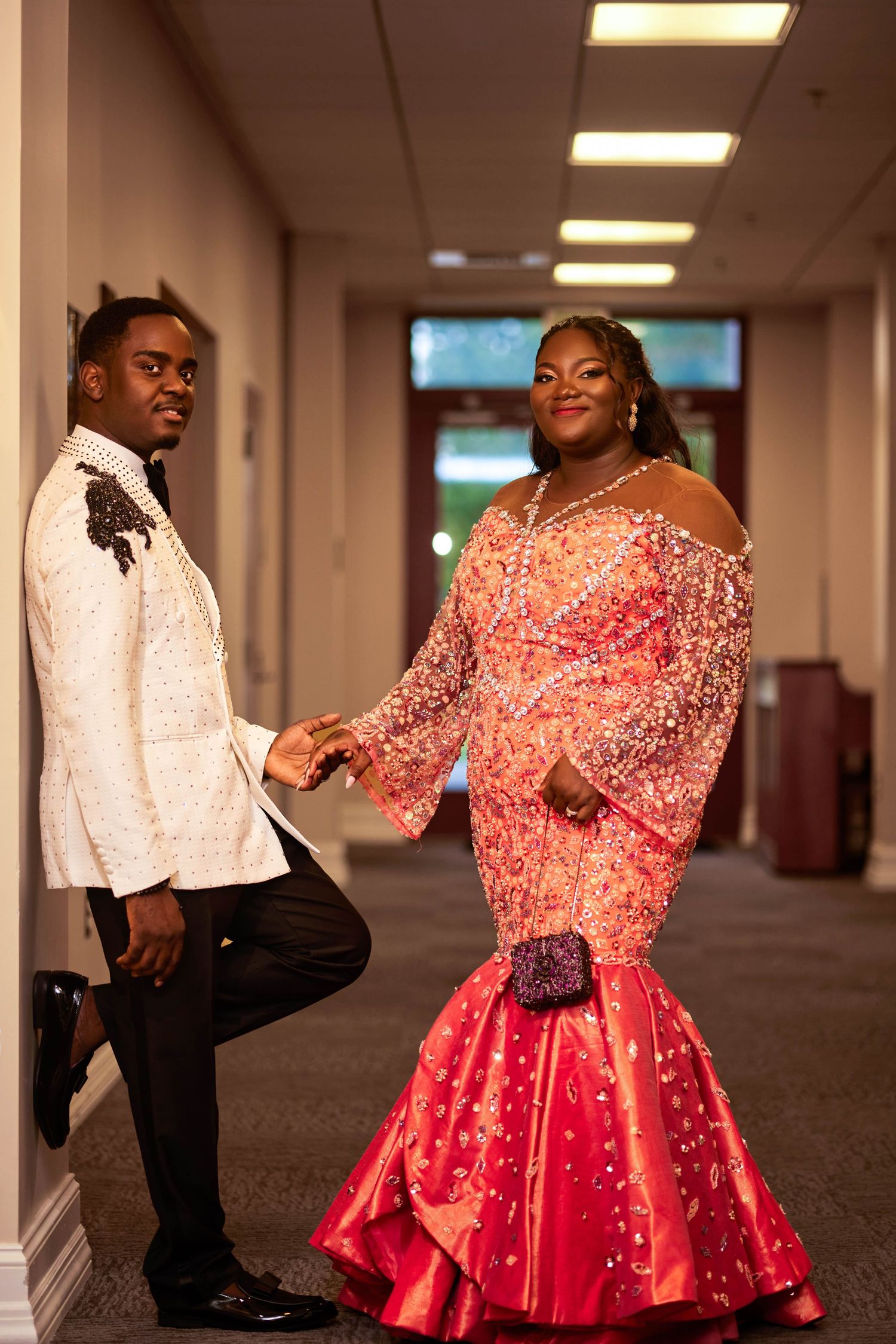 Elegant couple posing in hallway, man in white embroidered blazer, woman in sparkling red mermaid gown.