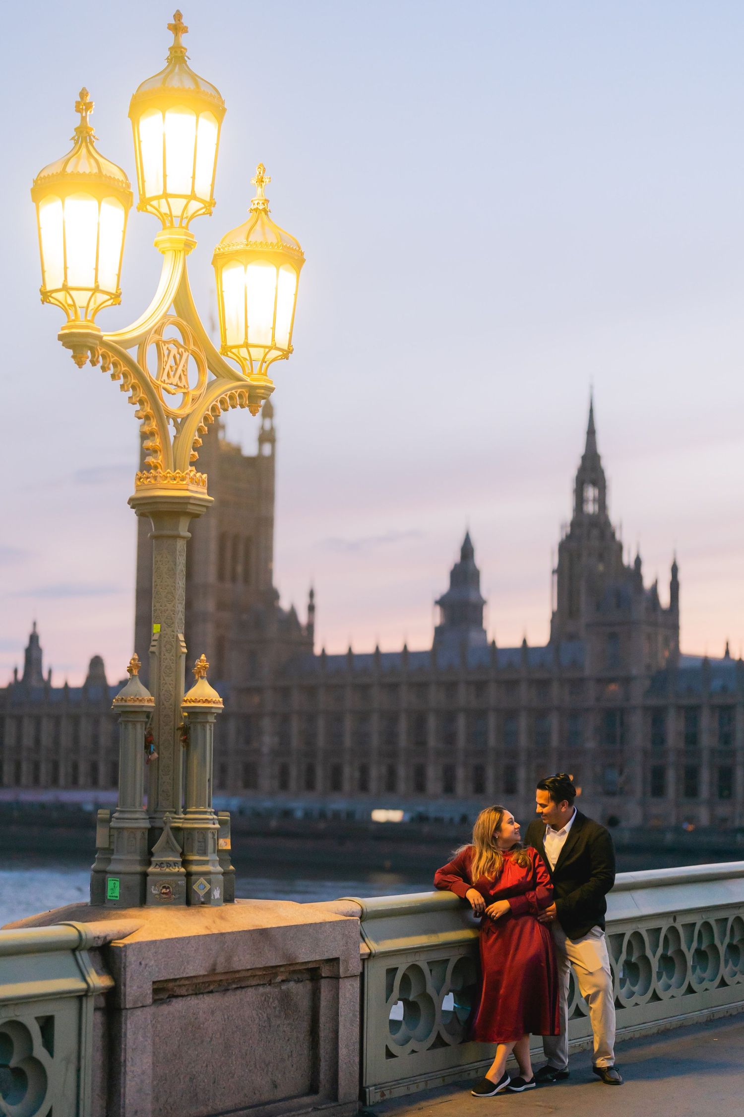 Couple sharing tender kiss before Tower Bridge London, pregnant woman in navy beaded gown, man in burgundy jacket.