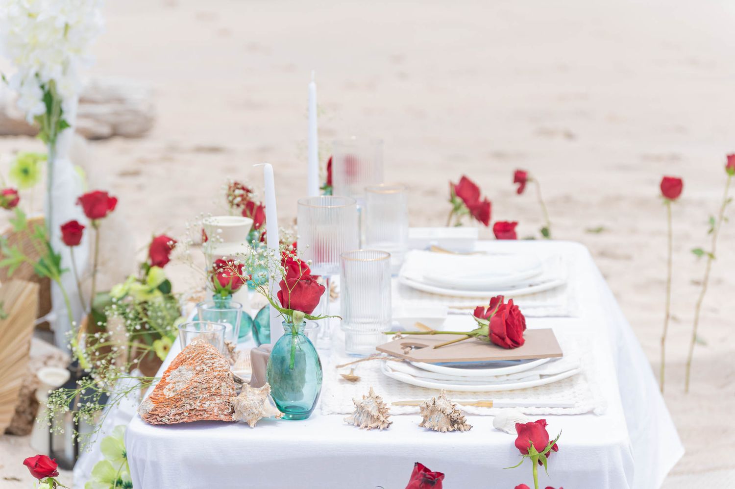 Elegant beach wedding table setting with red roses, white linens, candles, and seashells on sandy shore.
