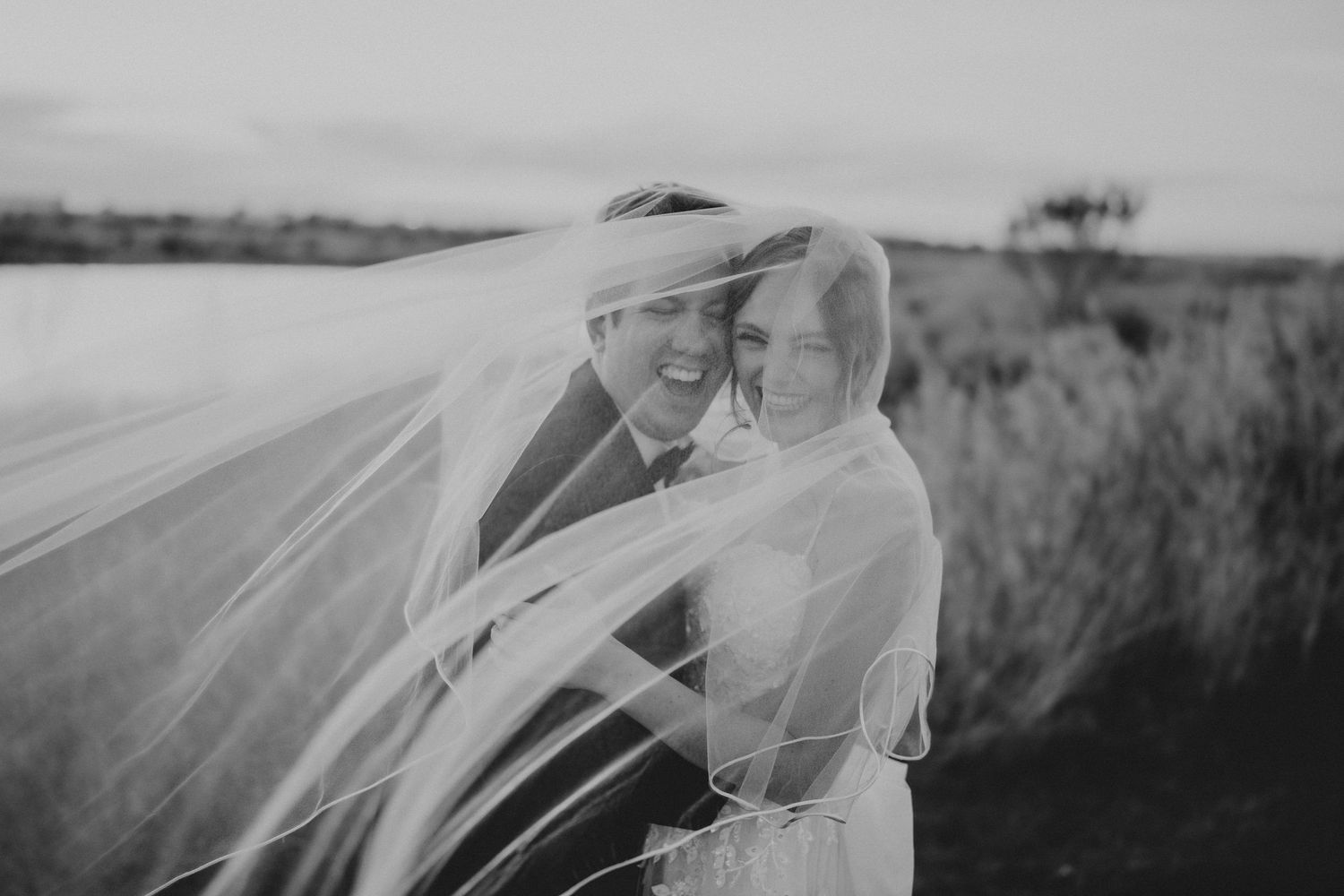Black and white wedding photo of smiling bride and groom wrapped in flowing veil outdoors.