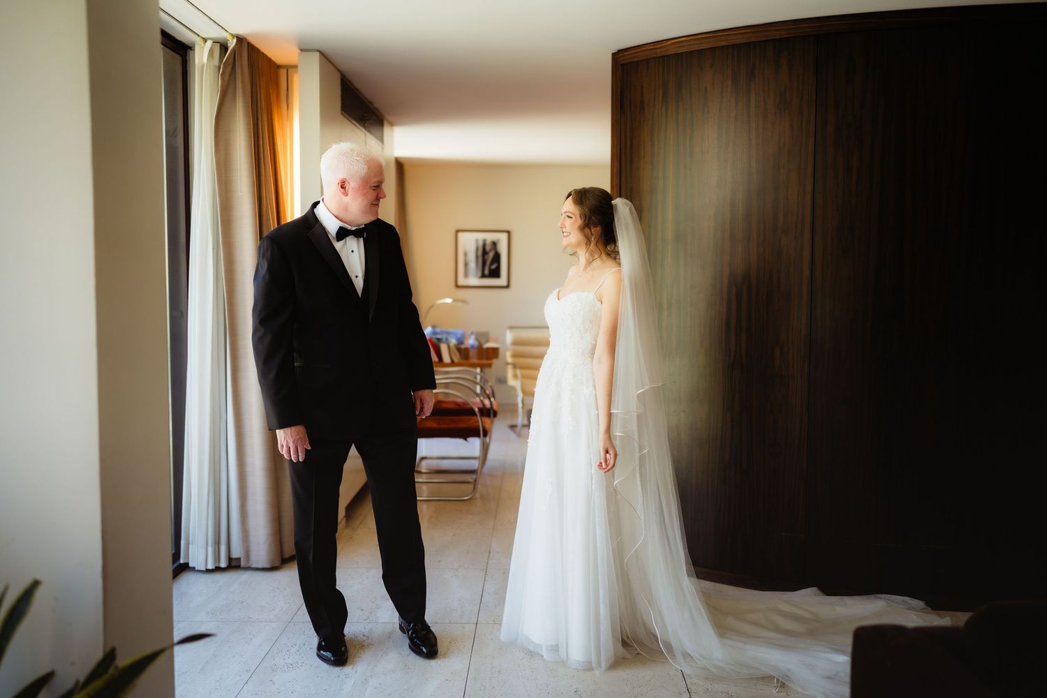 Father sees bride in wedding dress for the first time, emotional first look moment in elegant hotel room.