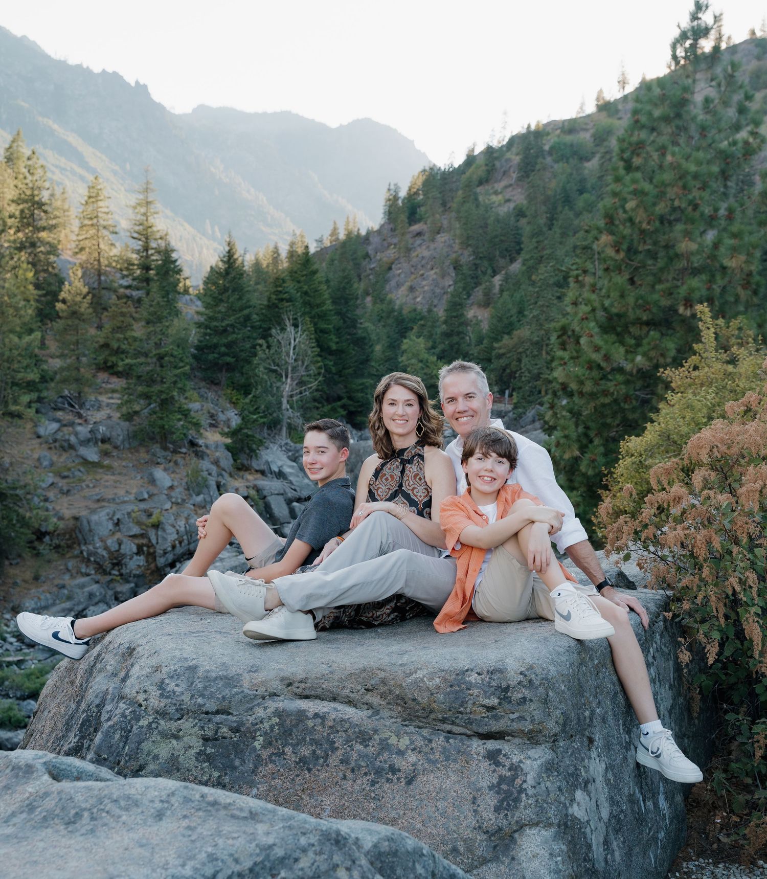 People relaxing on a large boulder in a scenic mountain landscape with evergreen trees and cliffs in the background.