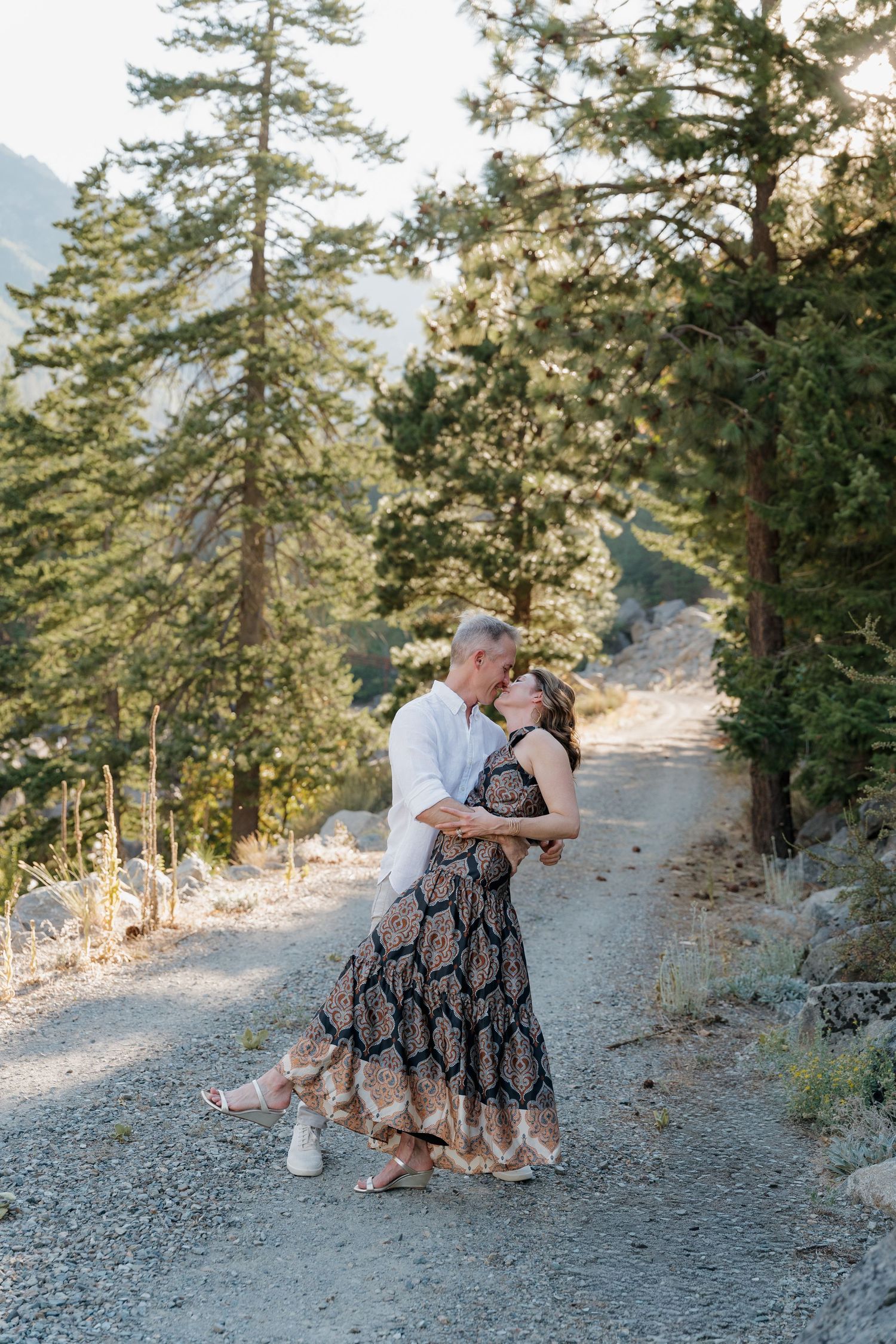 Couple shares an intimate moment on a rocky mountain trail surrounded by tall pine trees at sunset.