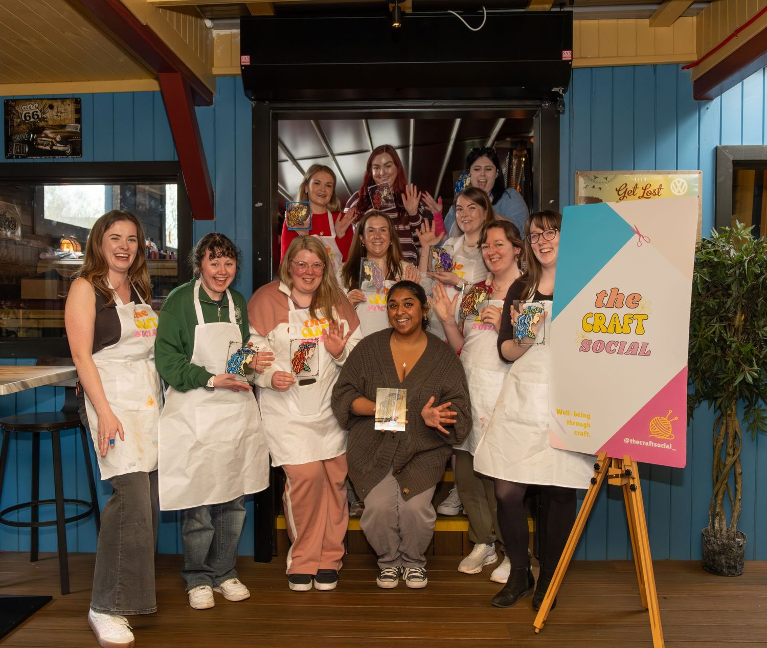 Group of women in aprons posing together at a craft social event near a colorful promotional banner.