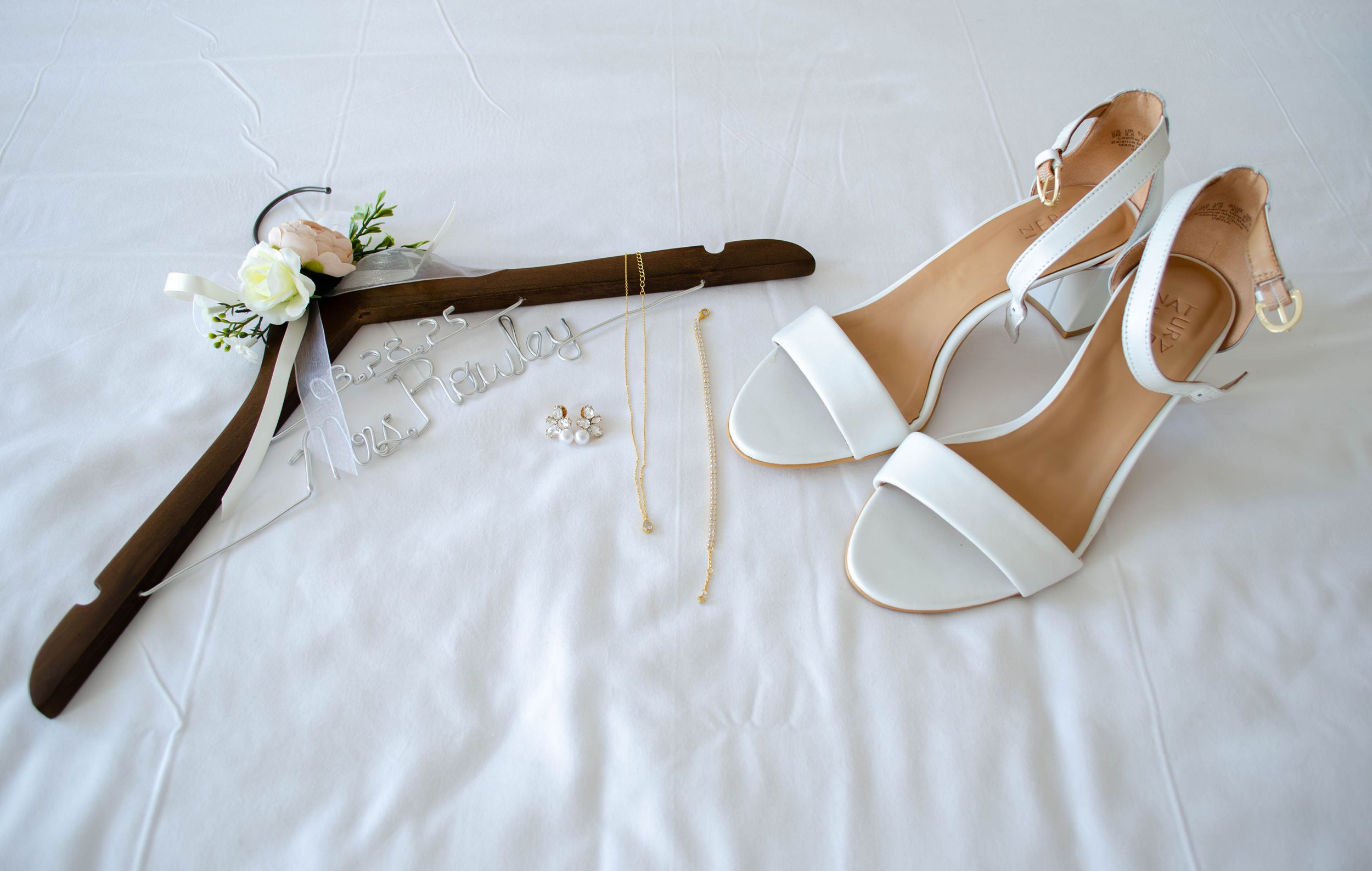 White bridal heels and floral wedding hanger with gold jewelry on white linen background.